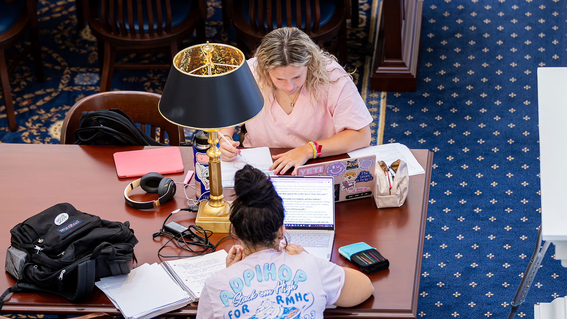 An overhead shot of two students studying together at a wooden table in a library. One student is writing by hand, while the other is looking at a laptop screen. The table holds a lamp, headphones, and notebooks. The floor is covered with a blue carpet with a star pattern.